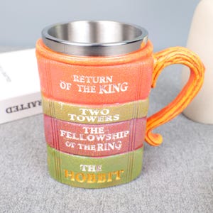 May include: A ceramic mug with a stainless steel interior, featuring a design of stacked books. The books are labeled "Return of the King," "Two Towers," "The Fellowship of the Ring," and "The Hobbit." The handle is orange and yellow.