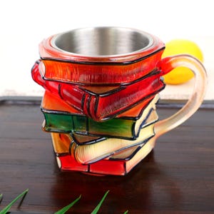 Puede incluir: Taza original con forma de pila de libros coloridos. La taza tiene un interior de acero inoxidable y un asa transparente. Los libros están pintados en tonos rojos, naranjas, verdes y amarillos.