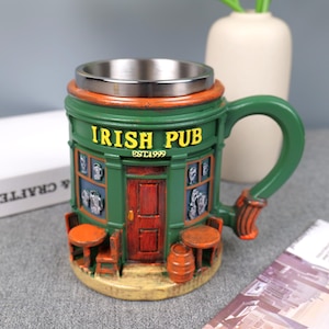 Puede incluir: Una jarra verde y naranja de Irish Pub con interior de acero inoxidable. La jarra presenta un diseño exterior detallado con las palabras "IRISH PUB EST1999". Tiene un asa y pequeñas mesas y sillas en el exterior.
