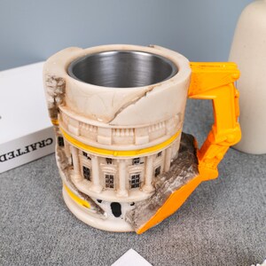 Könnte beinhalten: Eine neuartige Tasse mit Edelstahl-Innenraum und detailliertem Äußeren, das einem Gebäude ähnelt. Der Griff ist leuchtend orange und die Tasse hat ein gealtertes Aussehen. Die Tasse ist als Gebäude gestaltet.