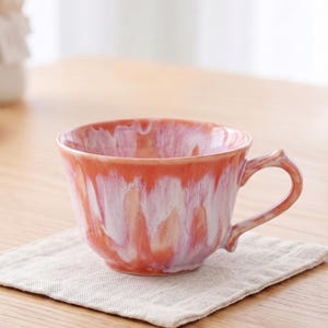 May include: A ceramic tea cup featuring a glossy glaze with a marbled pattern of coral orange, soft pink, and white. The cup has a wide, rounded bowl shape, a smooth rim, and a curved handle extending from the side, showcasing a fluid drip effect.