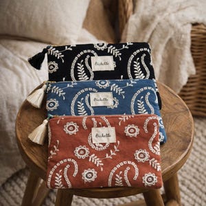 May include: Three rectangular zippered pouches in black, blue, and terracotta, each featuring white embroidered paisley and floral patterns. A small rectangular fabric label with the text Bichette is centered on each pouch, and a cream tassel is attached.