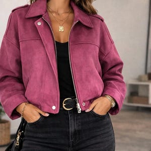 May include: A magenta suede jacket with a collared neckline and a silver zip. The jacket is paired with black jeans and a black belt with a gold buckle. A gold necklace and bracelet complete the outfit. The jacket's style is a classic biker jacket.
