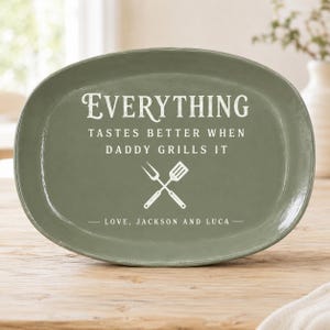 May include: An oval, olive-green serving platter with white text that reads "EVERYTHING TASTES BETTER WHEN DADDY GRILLS IT." Below the text is a crossed fork and spatula graphic, and the words "LOVE, JACKSON AND LUCA."