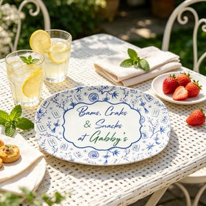 Puede incluir: Una bandeja de servicio ovalada blanca con un patr&oacute;n floral azul y fichas de mahjong. El centro muestra el texto "Bams, Craks & Snacks at Gabby's" en letra azul y verde. La bandeja est&aacute; colocada sobre una mesa de metal blanco con bebidas y fruta.
