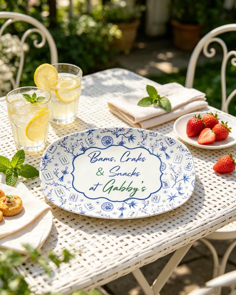 Puede incluir: Una bandeja de servicio ovalada blanca con un patr&oacute;n floral azul y fichas de mahjong. El centro muestra el texto "Bams, Craks & Snacks at Gabby's" en letra azul y verde. La bandeja est&aacute; colocada sobre una mesa de metal blanco con bebidas y fruta.