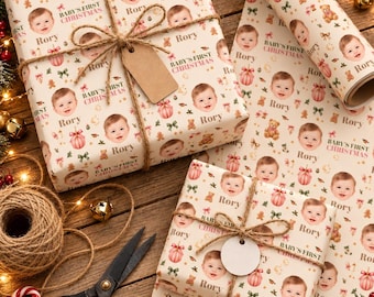 May include: Custom wrapping paper featuring a repeating pattern of a baby's face, the name Rory, teddy bears, pink ornaments, and the text Baby's First Christmas. Two gifts are wrapped in this paper, tied with twine and blank tags.