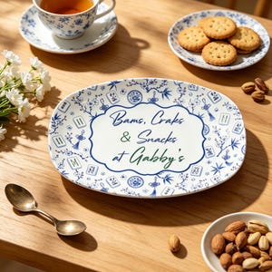 Puede incluir: Una bandeja de servicio ovalada de cer&aacute;mica blanca con un patr&oacute;n azul de flores y fichas de mahjong, con un borde decorativo central que contiene el texto Bams, Craks & Snacks at Gabby's en letras azules y verdes sobre una superficie de madera.