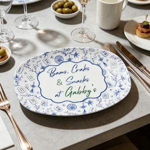 Puede incluir: Una bandeja de servicio ovalada de cer&aacute;mica blanca con un patr&oacute;n floral azul y blanco, borlas decorativas y motivos de fichas de mahjong. El centro muestra el texto en azul y verde: Bams, Craks & Snacks at Gabby's, en un borde festoneado.