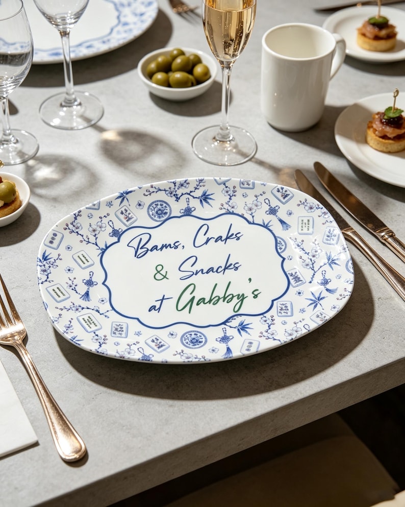 Puede incluir: Una bandeja de servicio ovalada de cer&aacute;mica blanca con un patr&oacute;n floral azul y blanco, borlas decorativas y motivos de fichas de mahjong. El centro muestra el texto en azul y verde: Bams, Craks & Snacks at Gabby's, en un borde festoneado.