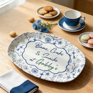 Puede incluir: Una bandeja de servicio ovalada de cer&aacute;mica con base blanca y un patr&oacute;n azul de flores y fichas de mahjong. El centro presenta un borde decorativo que encierra el texto "Bams, Craks & Snacks at Gabby's" escrito en letra cursiva azul oscuro y verde.