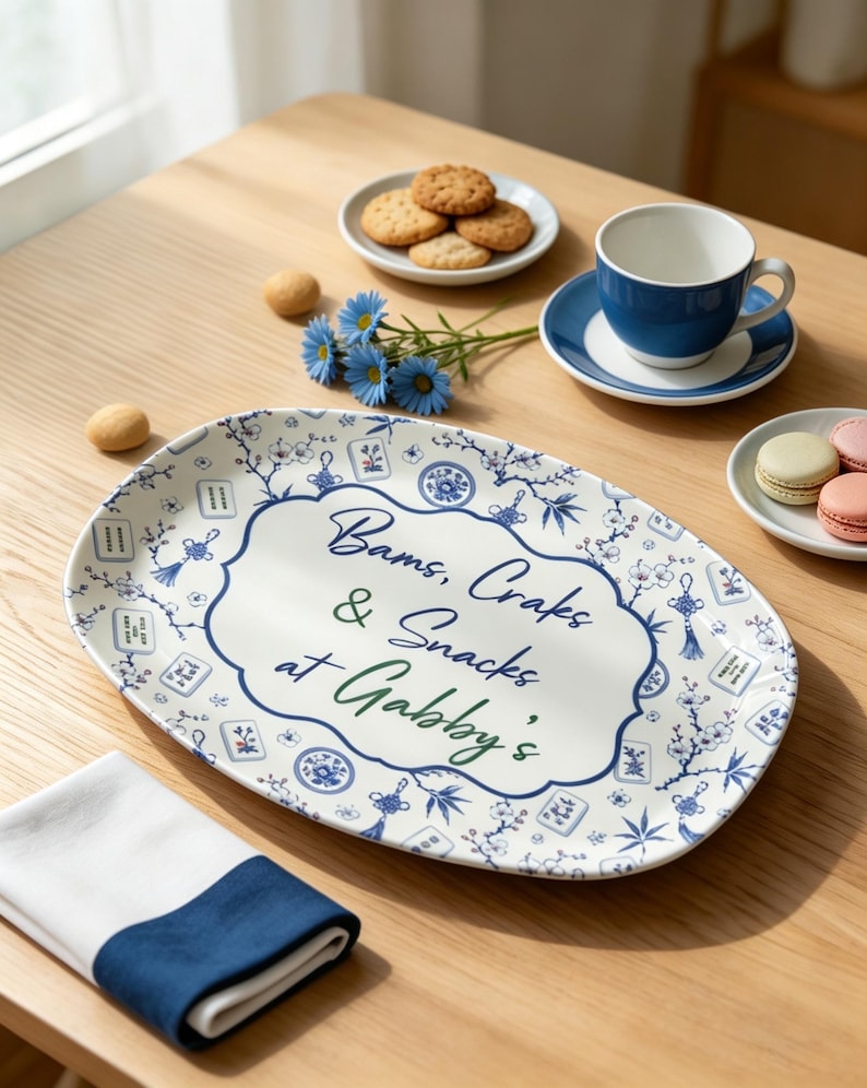 Puede incluir: Una bandeja de servicio ovalada de cer&aacute;mica con base blanca y un patr&oacute;n azul de flores y fichas de mahjong. El centro presenta un borde decorativo que encierra el texto "Bams, Craks & Snacks at Gabby's" escrito en letra cursiva azul oscuro y verde.