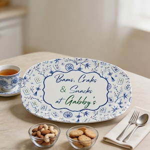 Puede incluir: Una bandeja de servir ovalada de cer&aacute;mica blanca con un patr&oacute;n floral azul y blanco y motivos de fichas de mahjong. El centro muestra el texto "Bams, Craks & Snacks at Gabby's" en letras azules y verdes sobre un fondo blanco.