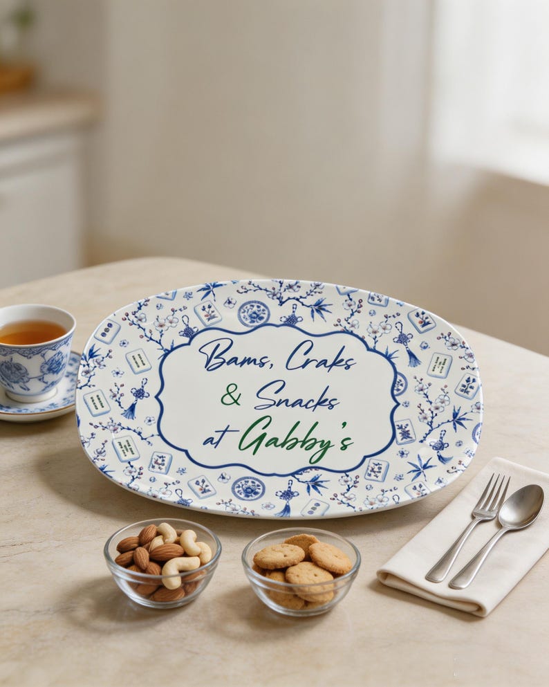 Puede incluir: Una bandeja de servir ovalada de cer&aacute;mica blanca con un patr&oacute;n floral azul y blanco y motivos de fichas de mahjong. El centro muestra el texto "Bams, Craks & Snacks at Gabby's" en letras azules y verdes sobre un fondo blanco.