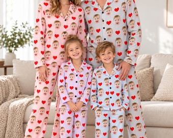 May include: Matching pajama sets featuring a repeating pattern of red hearts and a baby's face. The adult sets are in light pink and light gray, while the children's sets are in light purple and light blue, all with button-down tops.