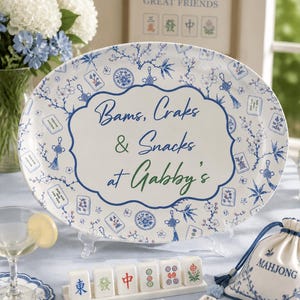 Puede incluir: Una bandeja de servir ovalada blanca con un patr&oacute;n de flores azules y fichas de mahjong. El centro presenta un borde decorativo con el texto "Bams, Craks & Snacks at Gabby's" en letra azul y verde. La bandeja se exhibe en un soporte.