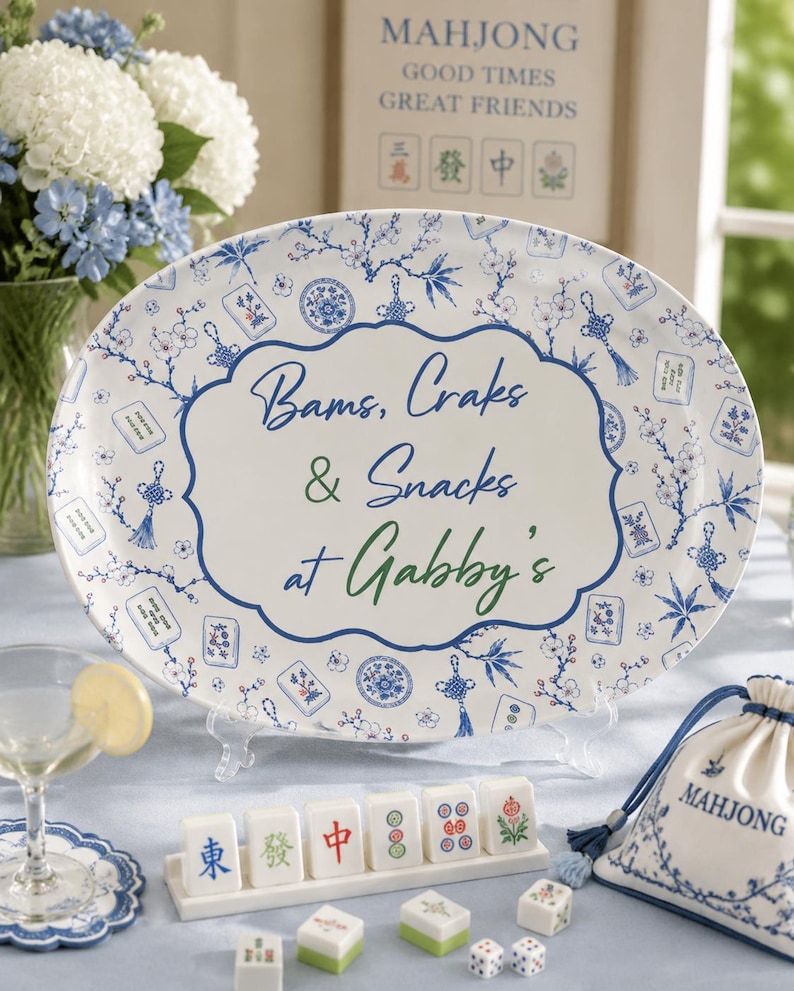 Puede incluir: Una bandeja de servir ovalada blanca con un patr&oacute;n de flores azules y fichas de mahjong. El centro presenta un borde decorativo con el texto "Bams, Craks & Snacks at Gabby's" en letra azul y verde. La bandeja se exhibe en un soporte.
