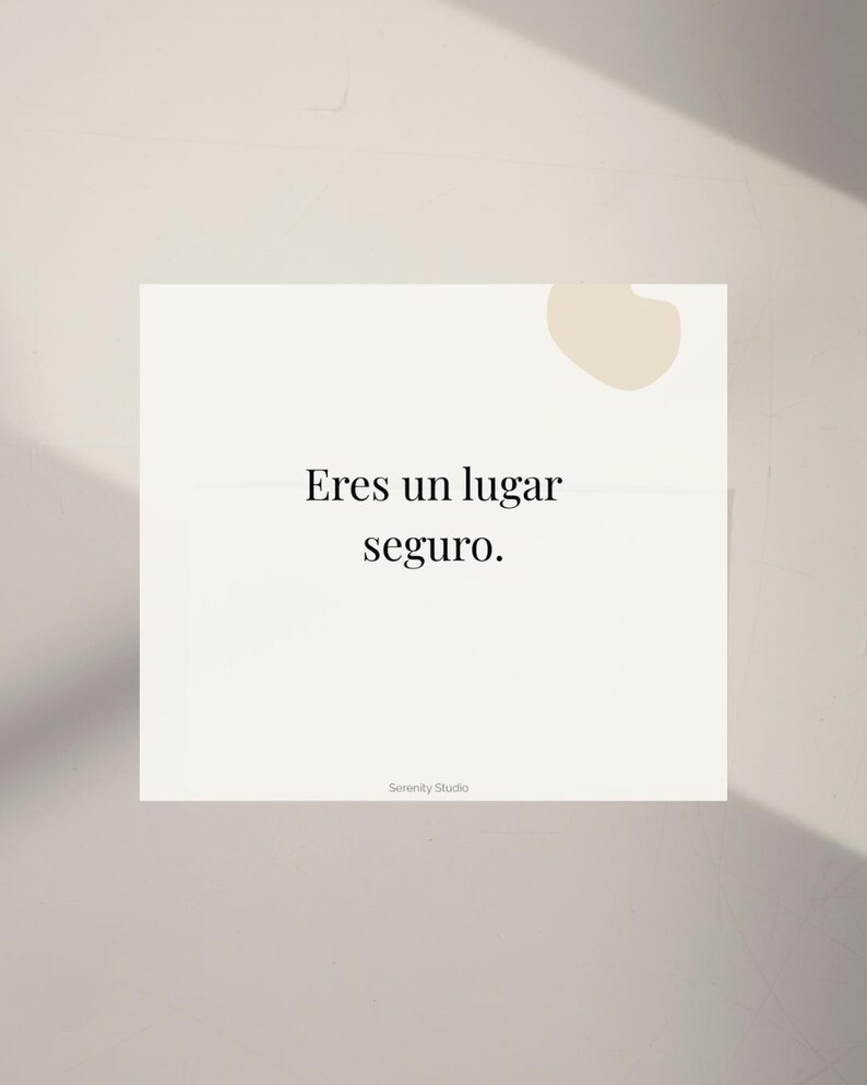 Op de afbeelding: Een witte kaart met de Spaanse tekst "Eres un lugar seguro" in een schreefletter. De kaart heeft een lichtbeige abstracte vorm in de rechterbovenhoek en de woorden "Serenity Studio" onderaan.