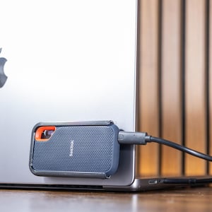 May include: A dark grey external hard drive connected to a silver laptop. The hard drive features an orange accent and the brand name "Sandisk". The laptop displays the Apple logo on its lid.