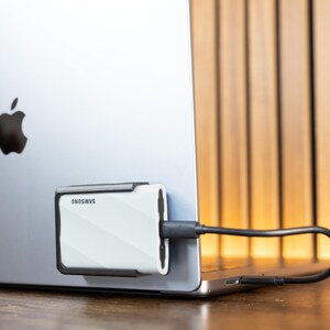 May include: A white Samsung external hard drive is connected to a silver laptop. The hard drive has a black border and a diagonal striped pattern. The laptop has the Apple logo on the back and a black cable is connected to the hard drive.