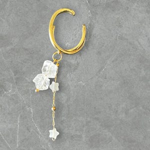 May include: A gold ear cuff with a curved design. The cuff has a dangling chain with clear, star-shaped and irregular charms. The jewellery is set against a grey background.