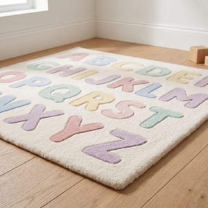 May include: A square area rug with a cream-colored base featuring the English alphabet in raised, tufted letters. The letters are arranged in rows and colored in soft pastel shades of pink, blue, yellow, green, and purple across the surface.