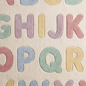May include: A plush area rug featuring a cream-colored background with raised, tufted letters of the alphabet in pastel shades of pink, blue, yellow, and green. The letters G, H, I, J, K, O, P, Q, R, and others are arranged in a grid pattern.