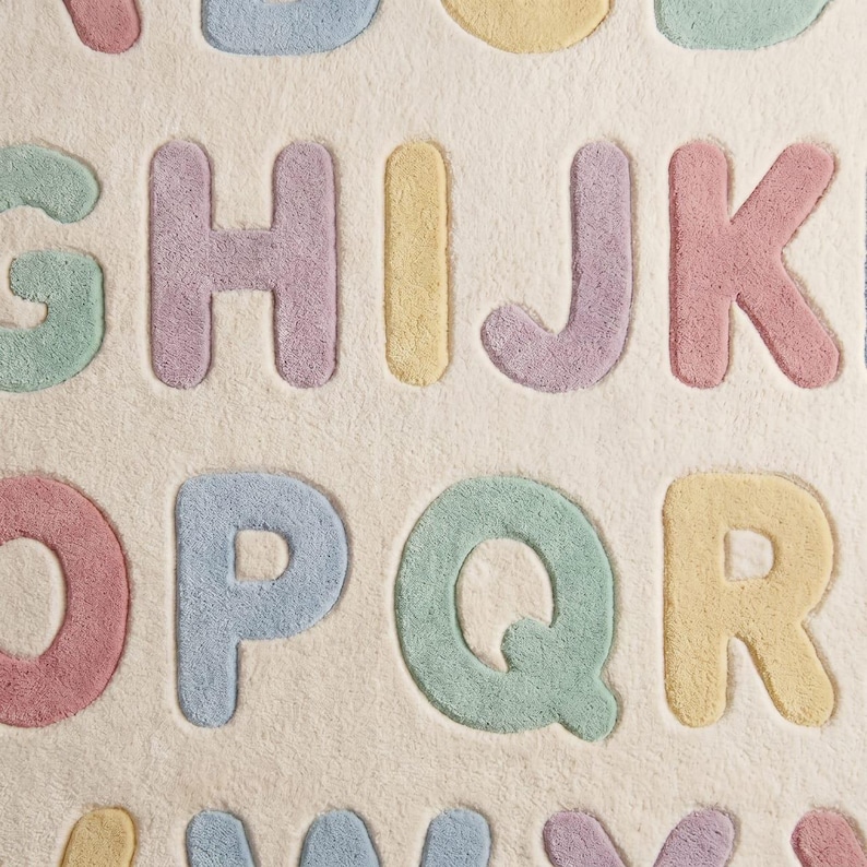 May include: A plush area rug featuring a cream-colored background with raised, tufted letters of the alphabet in pastel shades of pink, blue, yellow, and green. The letters G, H, I, J, K, O, P, Q, R, and others are arranged in a grid pattern.