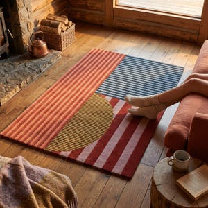 May include: A rectangular area rug featuring a geometric design with vertical stripes in shades of terracotta, dark blue, and mustard yellow, including a prominent semi-circular mustard yellow section set against a deep red and light pink striped background.
