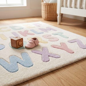 May include: A cream-colored area rug featuring the alphabet in raised, pastel-colored letters. A wooden block with the letter C and a single light pink leather moccasin are placed on the surface of the rug, which displays letters from A to Z.