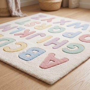 May include: A rectangular area rug featuring a cream-colored base with raised, tufted letters of the alphabet in pastel shades of pink, blue, yellow, and green. The letters are arranged in a repeating pattern across the soft, textured surface.
