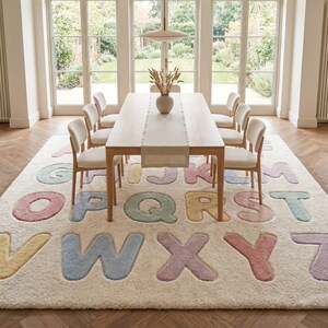 May include: A large rectangular area rug featuring a cream-colored background with a repeating pattern of raised, soft-textured alphabet letters in pastel shades of pink, blue, yellow, and green, covering the entire surface of the floor covering.