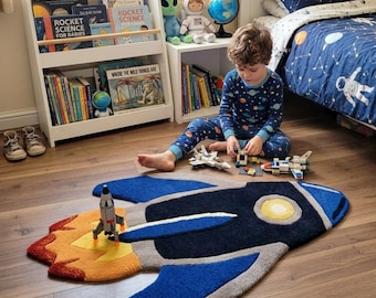 May include: A rocket-shaped area rug featuring a dark blue, light blue, and grey body with a yellow porthole and a bright orange and red flame design at the base, positioned on a wooden floor with a small toy rocket standing on the rug.