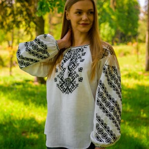 May include: White linen blouse with intricate black geometric embroidery on the bodice and sleeves. The long sleeves have a puffed design. The blouse is worn with dark blue jeans. The background is a blurred green outdoor scene.