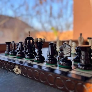 Puede incluir: Un juego de ajedrez de madera con piezas negras y blancas dispuestas sobre un tablero a cuadros. Las piezas de color marrón oscuro contrastan con los cuadros claros. El tablero está enmarcado con un borde decorativo.