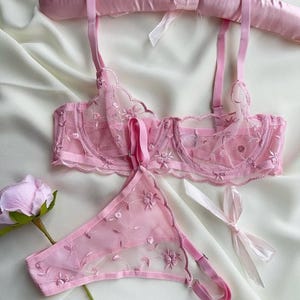 May include: A delicate pink lingerie set featuring a bra and matching thong, both adorned with floral embroidery. The bra has adjustable straps and underwire support. The set is displayed on a satin hanger, with a rose and ribbon nearby.