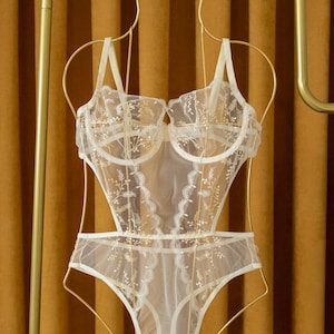 May include: A white sheer bodysuit featuring delicate gold floral embroidery, underwire cups, and scalloped lace trim along the vertical panels. The garment is constructed from translucent mesh fabric with adjustable shoulder straps and a high-cut leg.