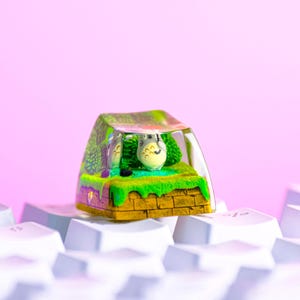 Peut inclure: Une touche de clavier mécanique en résine personnalisée représentant une créature grise miniature avec un parapluie noir sur une parcelle d'herbe vert vif avec une base en briques brunes, le tout dans un boîtier en résine transparent et brillant.