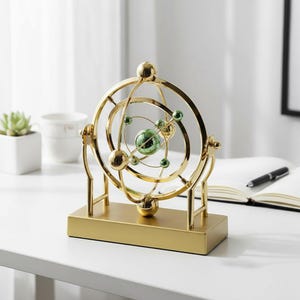 Könnte beinhalten: Goldfarbene Schreibtisch-Ornament im Orbitalstil mit rechteckigem Sockel. Das Design zeigt konzentrische Ringe und Kugeln in Gold und Grün, wodurch eine dynamische, wissenschaftliche Ästhetik entsteht. Das Ornament steht auf einer weißen Oberfläche.