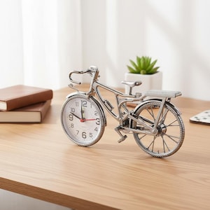 Puede incluir: Un reloj plateado con forma de bicicleta, con esfera blanca y números negros. El reloj tiene un segundero rojo y está colocado sobre una superficie de madera. Una pequeña planta en una maceta blanca está en el fondo.
