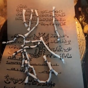 May include: A rectangular piece of tan paper featuring dark brown handwritten Arabic script and geometric symbols. Resting on top is a white cord tied into multiple small knots at regular intervals, creating a segmented, branch-like structure.