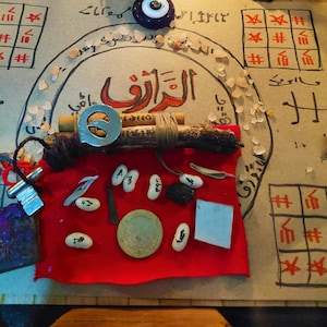 May include: A collection of esoteric objects displayed on a parchment background. A red cloth supports items like inscribed white beans, a small green disc, and a silver charm. The background includes handwritten text and symbols, alongside a blue and white eye-shaped bead.