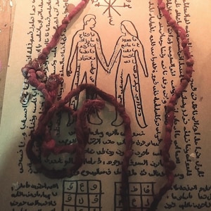 Può includere: Una collana di corda annodata rosso scuro con una spilla da balia in metallo, appoggiata su una pergamena color cuoio con un'illustrazione a inchiostro nero di due figure che si tengono per mano, circondate da scritte arabe e una griglia.