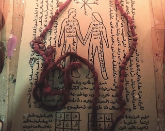 Puede incluir: Un collar de cordón anudado de color rojo oscuro con un imperdible de metal, sobre un pergamino color canela con una ilustración en tinta negra de dos figuras tomadas de la mano, rodeadas de escritura árabe y una cuadrícula.