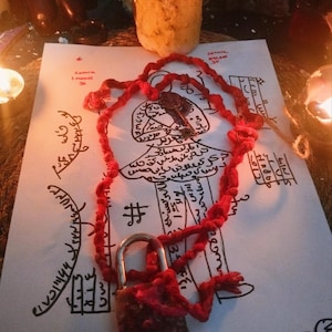 May include: A red cord necklace with a small padlock and a key pendant rests on a sheet of paper with handwritten text and illustrations. A lit candle is in the background, casting a warm glow.