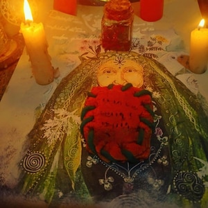 May include: A still-life arrangement featuring a red object with green accents, possibly a heart-shaped item, placed on a decorative cloth with a painted face. Lit candles and a jar with a cork lid are also present, creating a mystical ambiance.