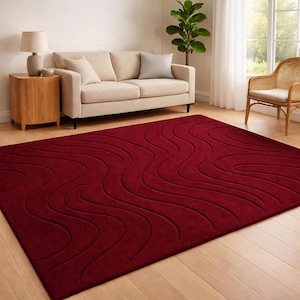 Könnte beinhalten: Ein großer, rechteckiger, weinroter Teppich mit einem wellenförmigen, strukturierten Muster. Der Teppich liegt auf einem Holzboden in einem Wohnzimmer, mit einem Sofa, einem Beistelltisch und einem Stuhl im Hintergrund.