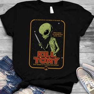 Könnte beinhalten: Schwarzes T-Shirt mit einem grünen Alien, das ein Messer hält, und dem Text "KILL TONY" in Rot und Gelb. Zusätzlicher Text lautet "The Undisputed No. 1 Live Podcast in the Galaxy" und "Airing Around the Universe from Austin, Texas!"