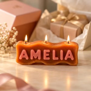 May include: A wavy, amber-coloured wax candle featuring three lit wicks and the name AMELIA embossed in raised, light pink capital letters across the front. The candle has a smooth, moulded texture and is positioned in a gift-ready setting.