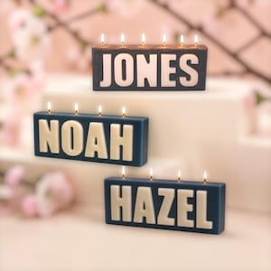 May include: Three rectangular block candles with lit wicks. The top candle is dark grey with the white text JONES. The middle candle is blue with the white text NOAH. The bottom candle is blue with the white text HAZEL. Each candle features raised lettering.